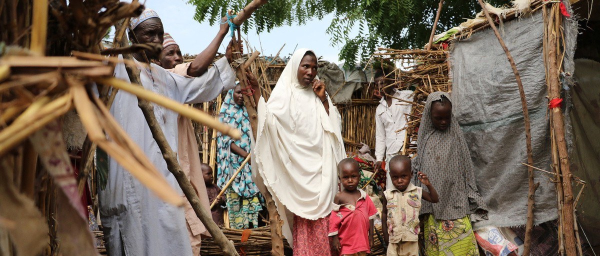 Women, men and children stand outside makeshift shelters of sticks, straw and plastic sheeting